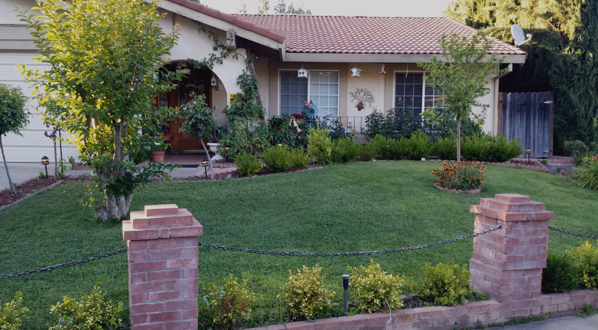 Front view of a single-story house with a well-maintained green lawn, small trees, shrubs, and flower beds. The house has a tiled roof, two large windows, a wooden front door, and a small porch area with decorative plants. A low brick fence with a chain runs along the front of the property.