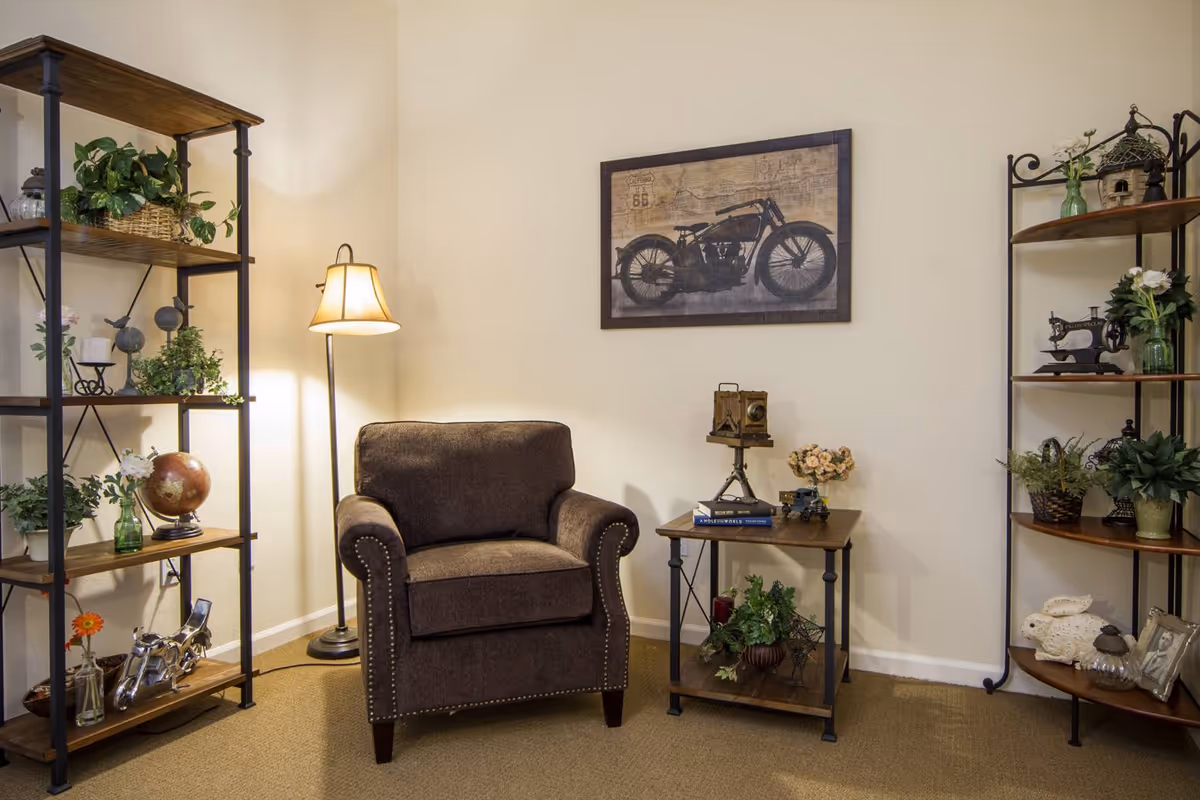 Cozy sitting area with a brown armchair, floor lamp, side table and decorative shelving against a neutral wall.