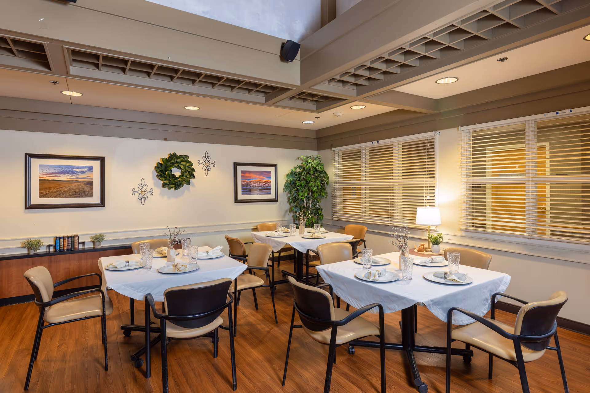 Well-lit dining room with multiple tables set for meals, chairs, framed artwork, plants, and window blinds.