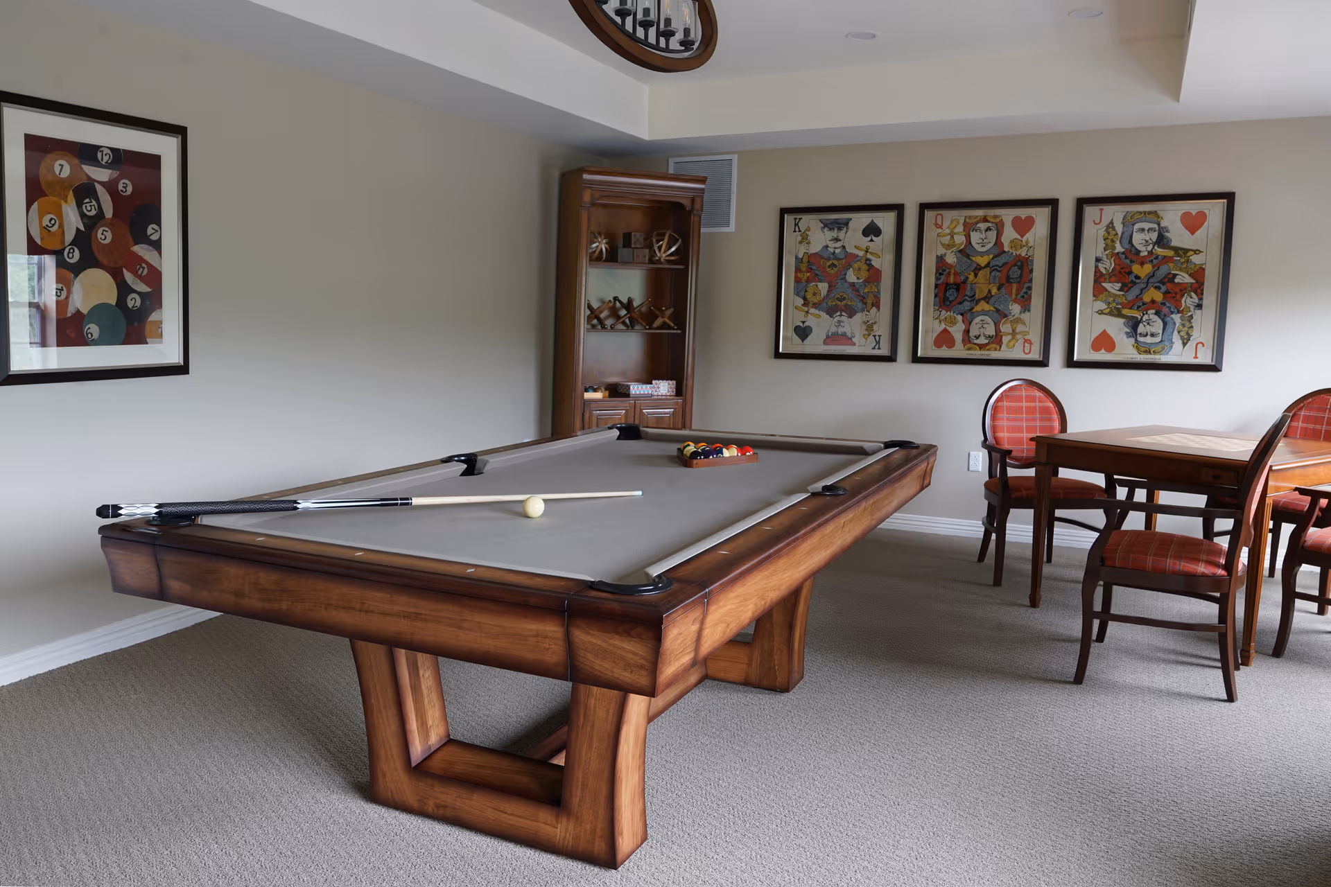 A game room featuring a wooden pool table with a cue stick and billiard balls arranged on the table. The room has beige walls decorated with framed artwork of playing cards and billiard balls. There is a wooden cabinet with shelves holding various items and a wooden table with four red cushioned chairs in the corner.
