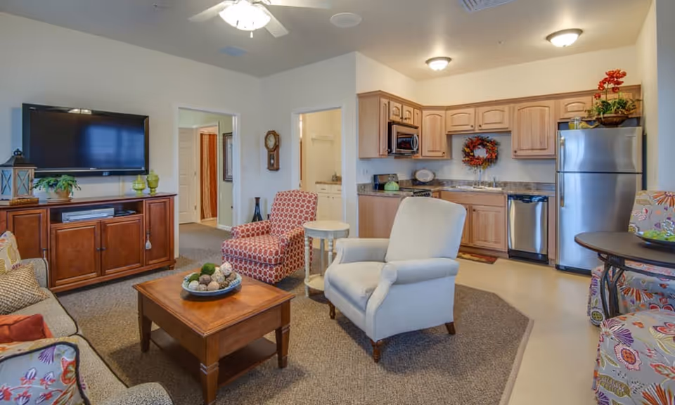 Cozy open living room with armchairs, a coffee table, wall-mounted TV and an adjacent kitchenette with wooden cabinets and stainless steel appliances.
