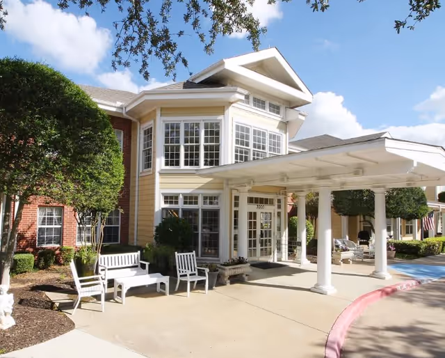 Front entrance of a two-story yellow-and-brick senior living building with a covered porte-cochere, white outdoor benches, and landscaping.