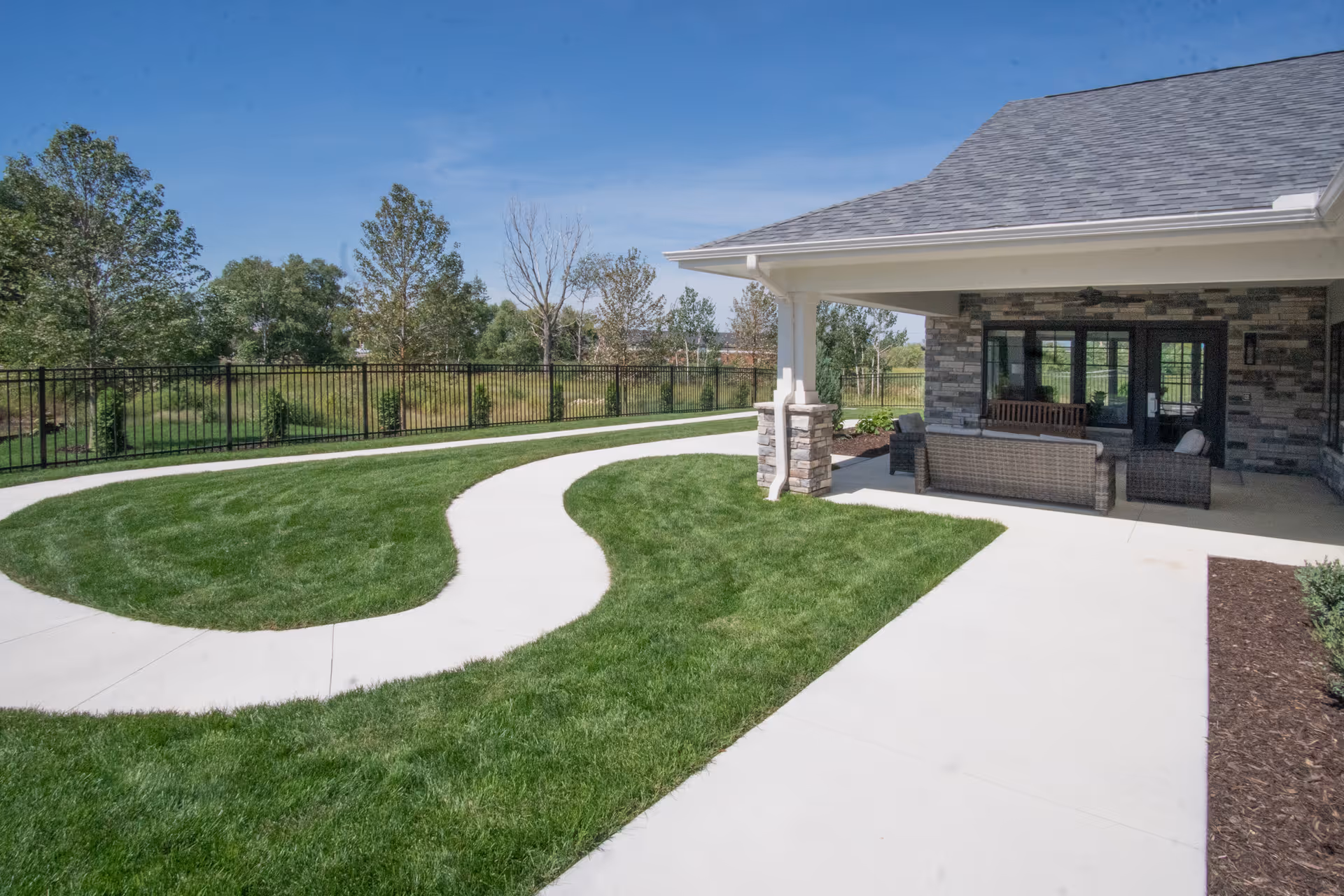 Covered patio with wicker seating next to a curving concrete walkway and manicured lawn in a landscaped outdoor area.