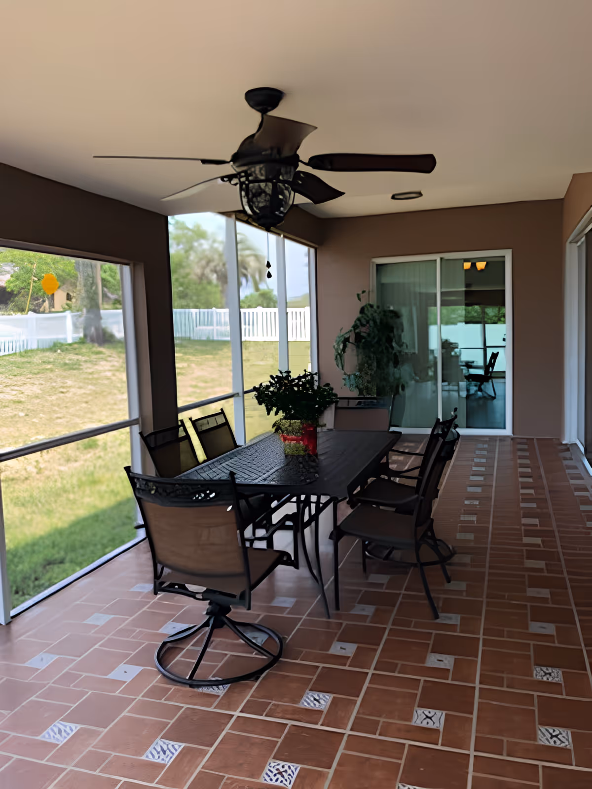 A covered outdoor patio area with a ceiling fan, a rectangular metal table, and six chairs. The floor is tiled with a brown and beige pattern. There are potted plants on the table and near the sliding glass doors that lead inside. The patio is enclosed with mesh screens, and a grassy yard with a white fence is visible outside.