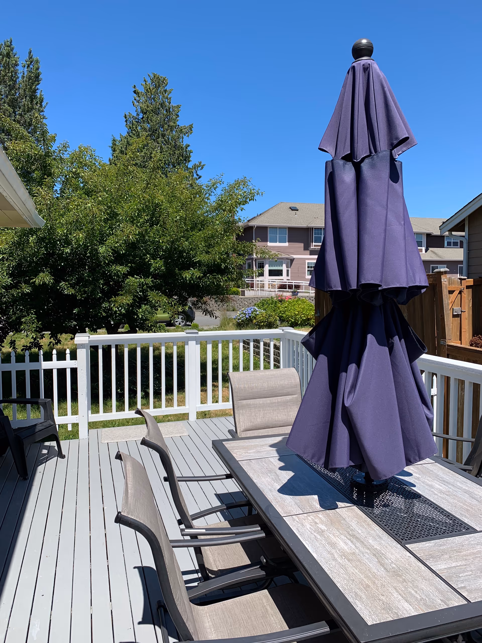 Outdoor wooden deck with a dining table, several chairs and a folded purple patio umbrella overlooking a white fence and neighboring houses.