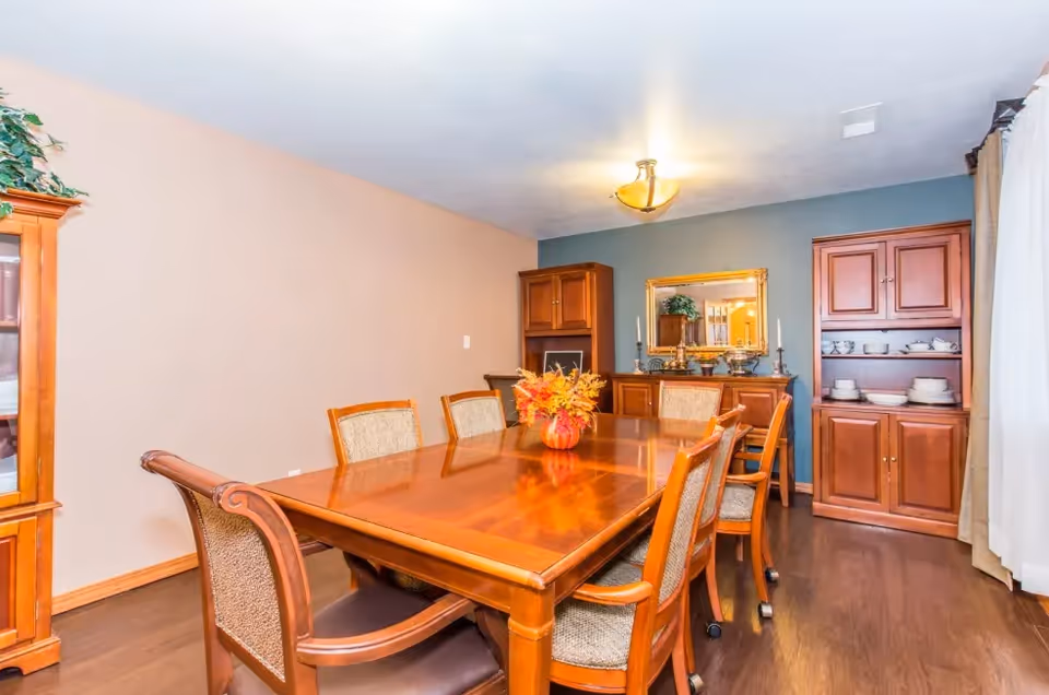 A dining room with a polished wooden table surrounded by eight cushioned chairs. The table has a centerpiece with autumn leaves in a small vase. The room features wooden cabinets filled with dishes, a large mirror on the wall, and a ceiling light fixture. The walls are painted in beige and blue tones, and there is a window with white curtains on the right side.