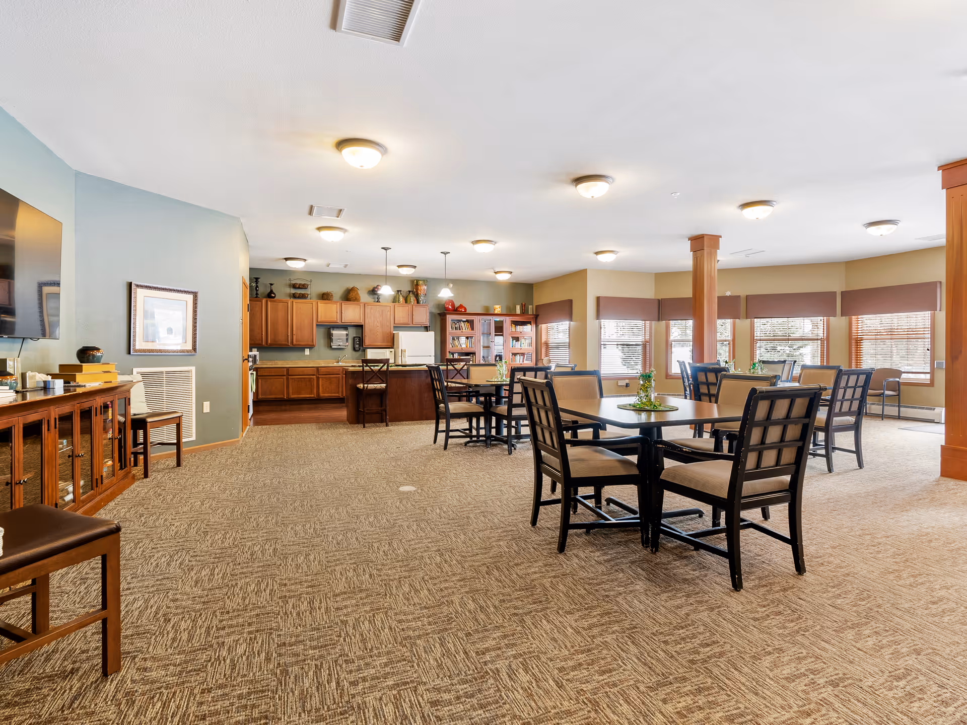 A spacious senior living common area with multiple tables and chairs arranged for dining or socializing. The room features large windows with blinds, carpeted floors, and a kitchen area with wooden cabinets and a refrigerator in the background. The walls are painted in soft tones, and the ceiling has multiple light fixtures providing bright illumination.