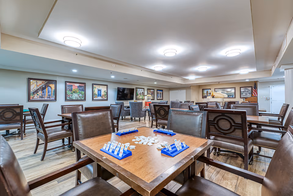 A spacious senior living common area with multiple tables and chairs arranged for group activities. One table in the foreground has a game set up with tiles and racks. The room features wood flooring, framed artwork on the walls, a flat-screen TV, and a white grand piano in the background. The ceiling has recessed lighting fixtures providing bright illumination.