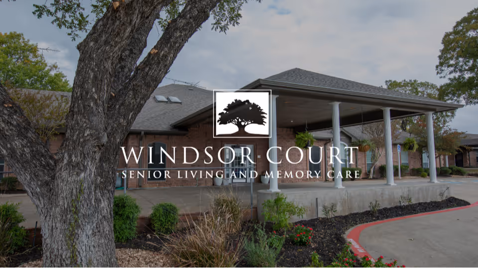 Front entrance of Windsor Court Senior Living building showing a covered drop-off with columns, landscaping, and a large tree in the foreground.