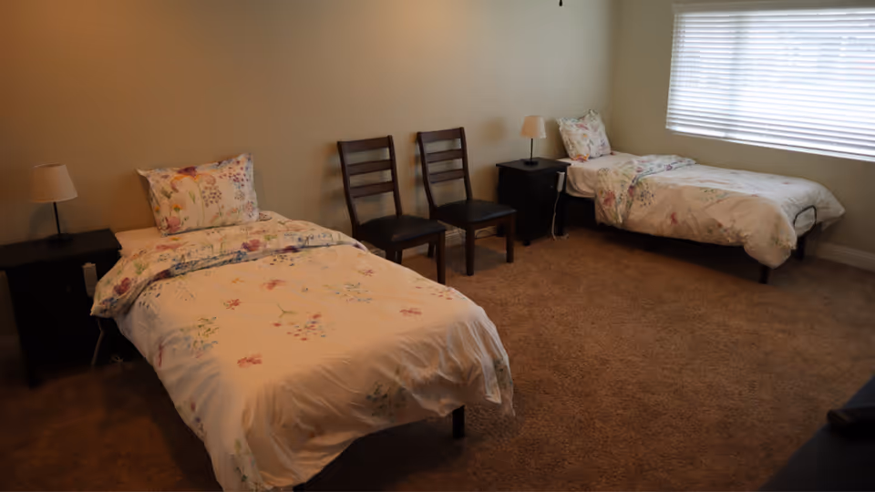 A bedroom with two single beds, each with floral bedding and a pillow. Between the beds are two wooden chairs and two black nightstands, each with a small lamp. A large window with blinds is on the right wall, letting in natural light. The room has beige carpet and light-colored walls.