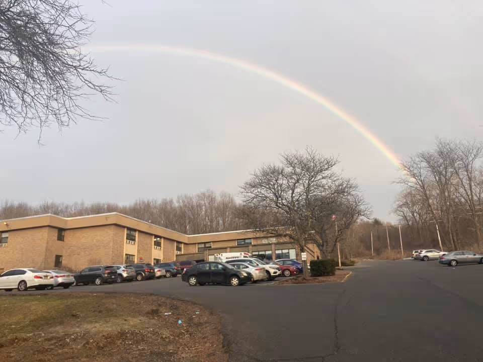 Brick two-story rehabilitation building with a parked cars in front, bare trees, and a rainbow arching across the sky.