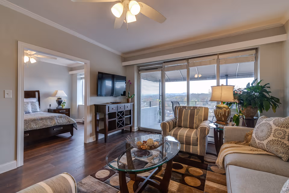 A cozy living room with a striped armchair, a glass coffee table, a beige sofa with patterned pillows, and a side table with a lamp and plants. A flat-screen TV is mounted on the wall above a wooden console. Large sliding glass doors lead to a balcony with outdoor seating and a scenic view. A bedroom with a bed, nightstand, and lamp is visible through an open doorway.