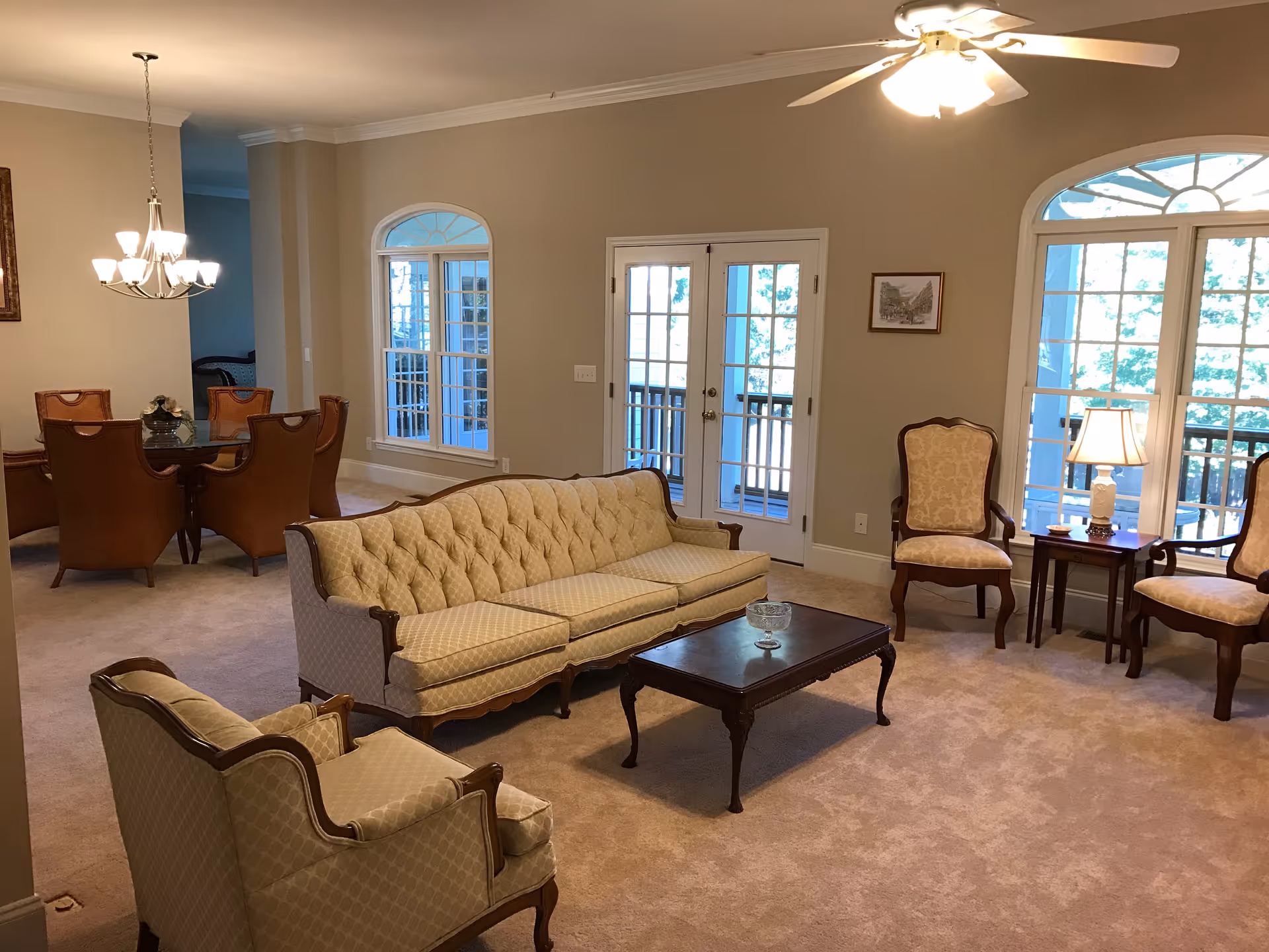 A cozy living room with beige carpet and walls, featuring a tufted beige sofa, two matching armchairs, a wooden coffee table, and a side table with a lamp. The room has large windows and French doors leading to a balcony, with a ceiling fan and light fixture overhead. In the background, there is a dining area with a glass-top table and six brown leather chairs under a chandelier.