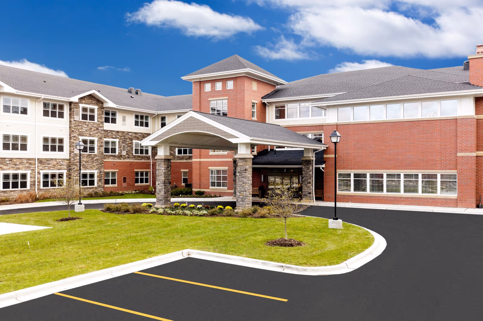 Exterior view of Clarendale of Algonquin senior living facility showing a multi-story building with a covered entrance, brick and stone facade, large windows, a well-maintained lawn, and a parking area under a bright blue sky with some clouds.
