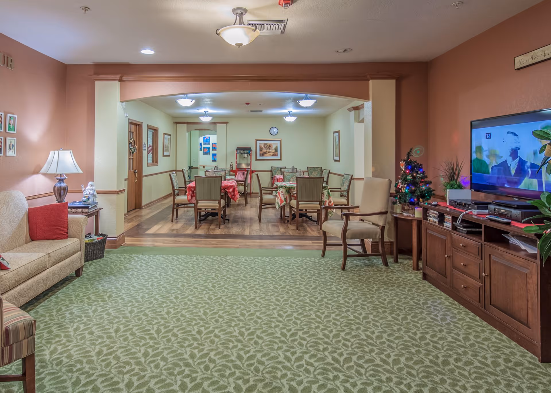A cozy senior living facility common area with a green patterned carpet and a beige couch with red pillows on the left. A wooden side table with a lamp and decorative items is next to the couch. In the background, there is a dining area with several tables covered with red and green tablecloths and surrounded by chairs. On the right side, there is a wooden entertainment unit with a flat-screen TV showing a black and white image, a small decorated Christmas tree, and some plants. The walls are painted in warm tones with framed pictures and soft lighting fixtures on the ceiling.