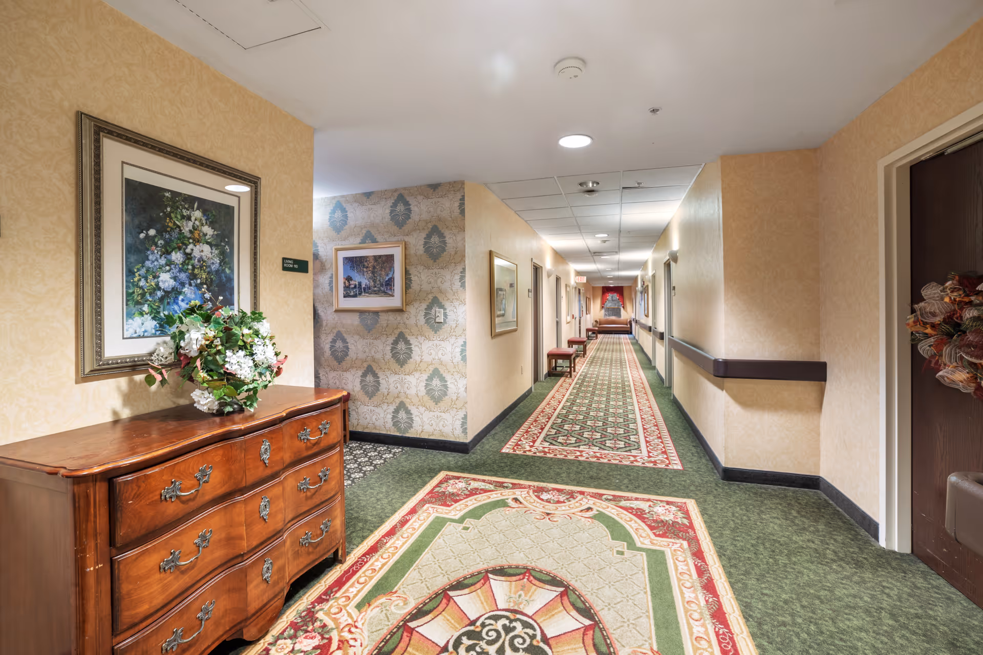 Carpeted interior hallway with patterned runners, framed artwork, a wooden dresser topped with flowers, and doors along the corridor.