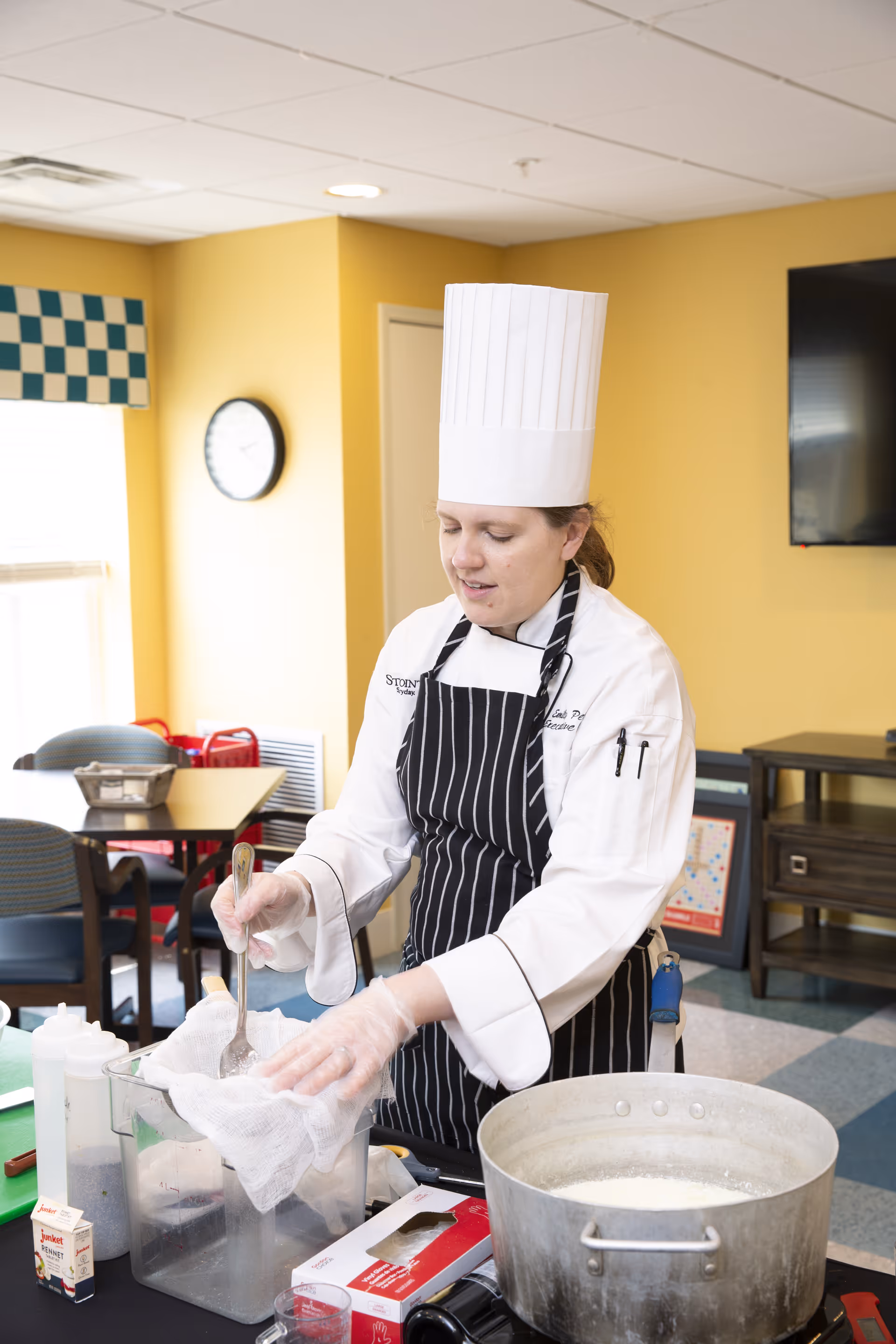 A chef wearing a white chef's hat, white coat, and black striped apron is preparing food in a kitchen or dining area. The chef is holding a spoon and a cloth over a clear container. There is a large metal pot on the table filled with a white liquid, along with various kitchen items and ingredients. The background shows a yellow wall, a clock, tables, chairs, and a television mounted on the wall.