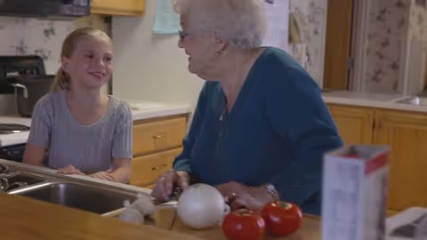 An elderly woman and a young girl smiling and talking at a kitchen counter with tomatoes and an onion visible.