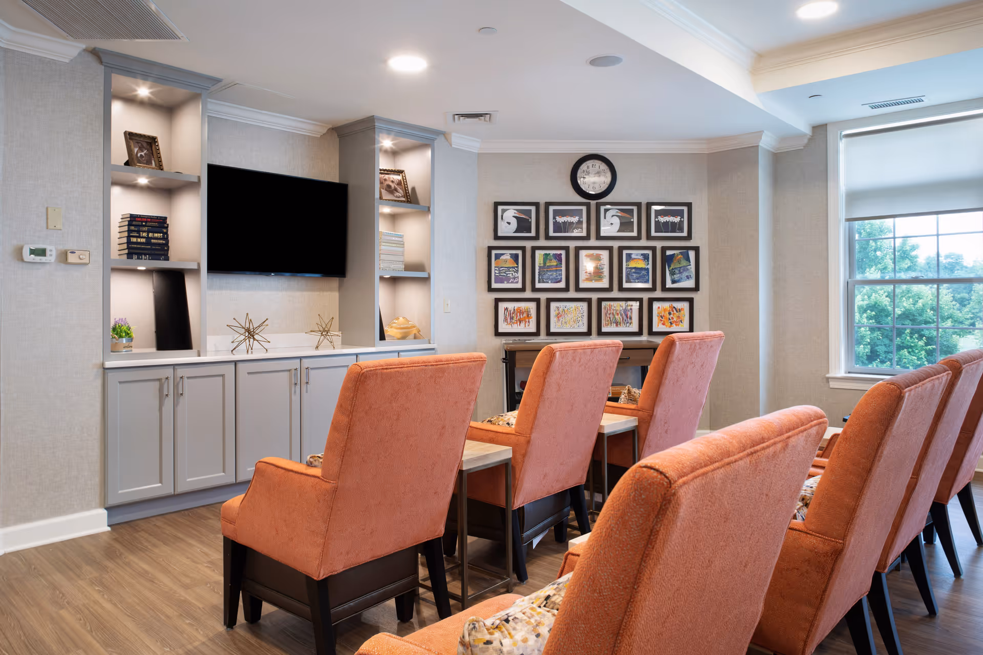 A cozy living room area with six coral-colored armchairs arranged in two rows facing a wall-mounted flat screen TV. The TV is flanked by built-in shelves holding books, decorative items, and framed pictures. On the opposite wall, there is a collection of framed artwork and a clock above them. A large window with a roller shade lets in natural light, and the room has wood flooring and neutral-colored walls.
