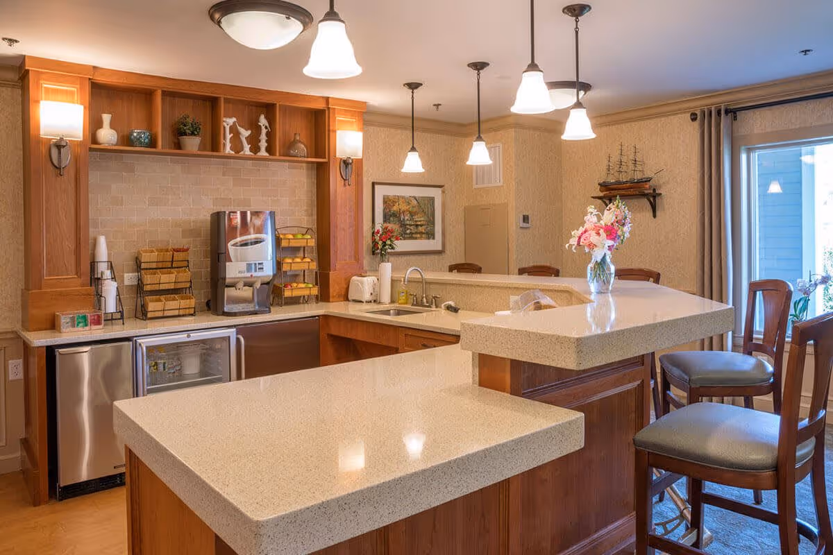 A well-lit kitchen area with a large beige countertop island and wooden cabinets. The kitchen features a coffee machine, a small refrigerator, a sink, and various decorative items including flowers in a vase and small sculptures on shelves. There are pendant lights hanging from the ceiling and wooden chairs with cushioned seats around the counter. A window with curtains is visible in the background.