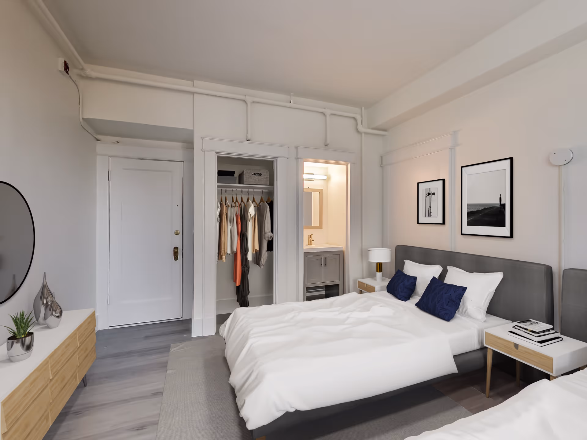 Modern bedroom with two beds, nightstands, framed artwork, an open closet with hanging clothes, and a small vanity.