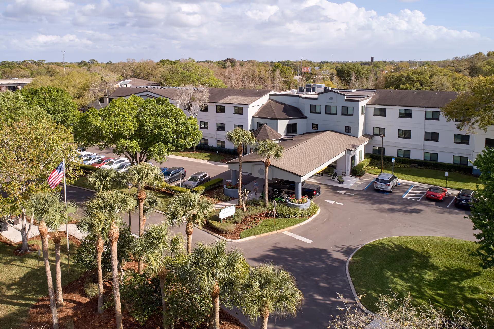 Aerial view of Renaissance North Tampa senior living facility showing a three-story building with a covered entrance, surrounded by palm trees, greenery, and a parking lot with several cars. An American flag is visible near the entrance.