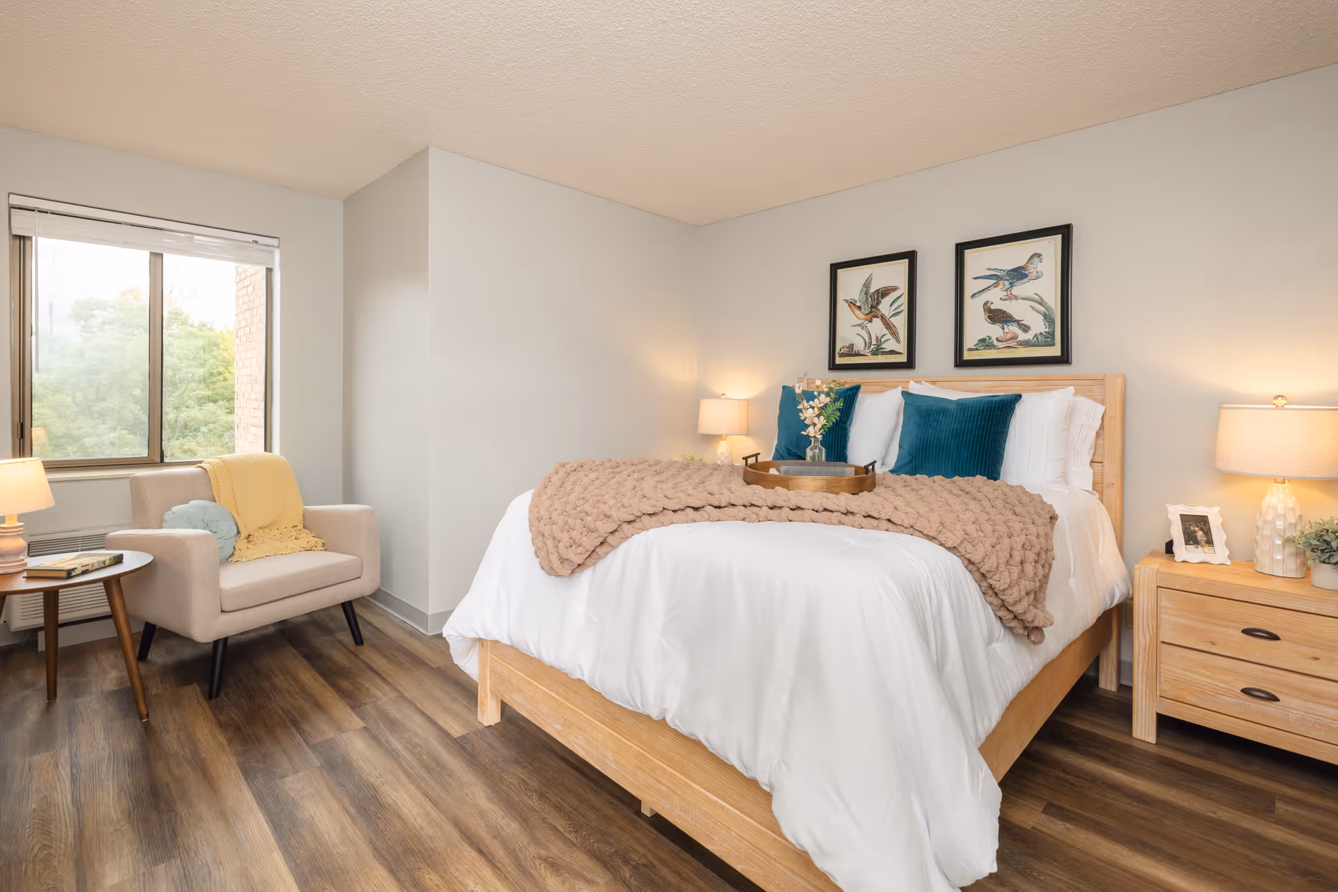 Well-lit bedroom with a made bed, nightstands and lamps, an armchair by a window, and framed bird prints above the bed.