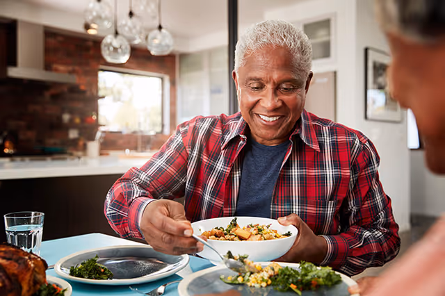 An elderly man with gray hair wearing a red plaid shirt is smiling and serving food from a bowl onto his plate at a dining table in a modern kitchen setting.