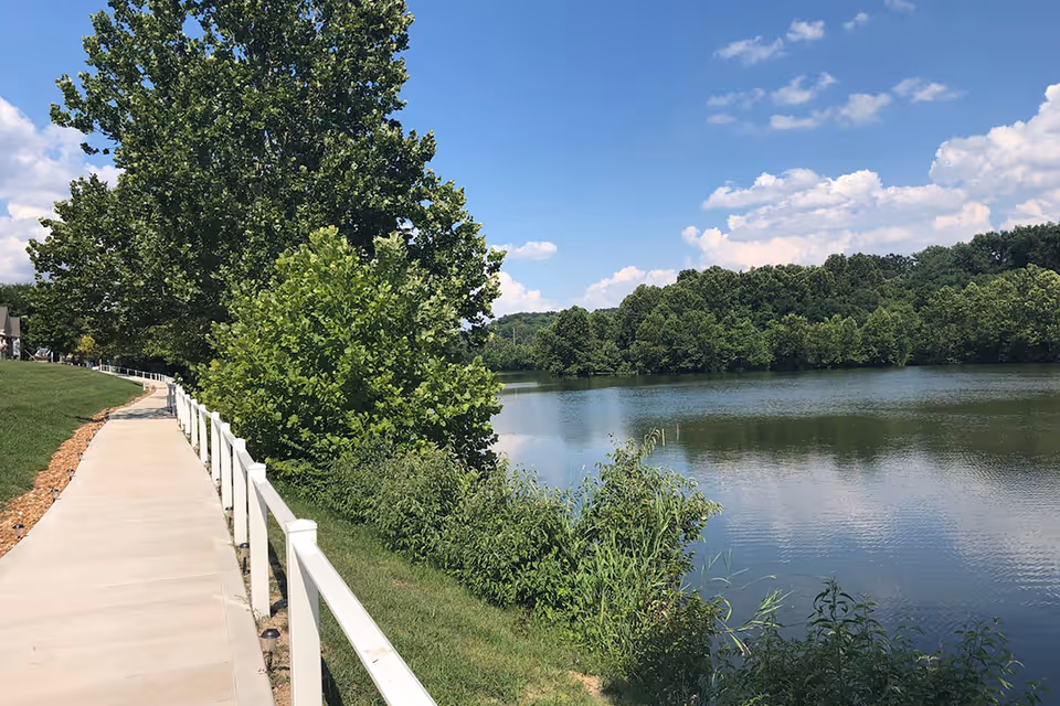 A paved walkway with a white railing runs alongside a calm lake surrounded by green trees under a blue sky with scattered clouds.