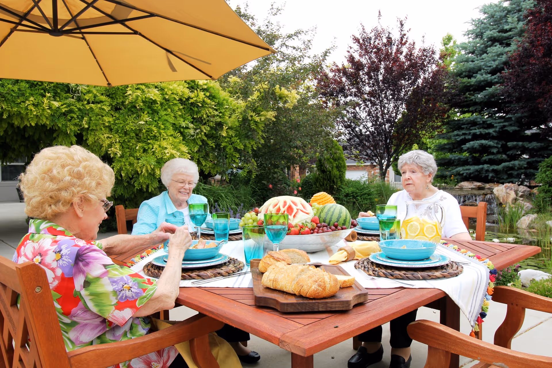 Three elderly women sitting around a wooden outdoor table under a large yellow umbrella, enjoying a meal with plates, glasses, a pitcher of lemonade, bread, and a bowl of assorted fruits. The setting is a lush garden with green trees and shrubs in the background.
