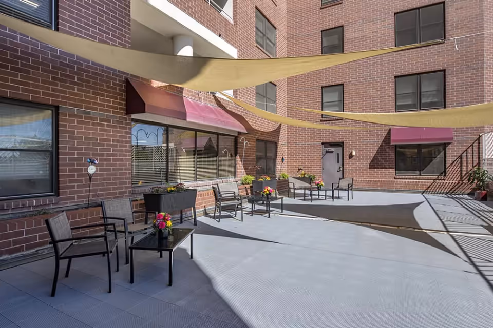 Outdoor patio area at Brookdale Meridian Englewood with several chairs and tables arranged along a brick building wall. There are flower pots on the tables and planters with flowers along the wall. Beige shade sails are stretched overhead providing partial shade.