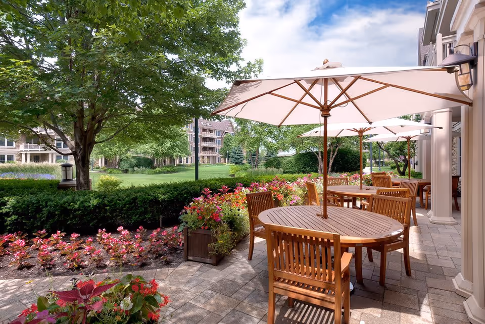 Outdoor patio area with wooden tables and chairs under large white umbrellas, surrounded by colorful flowers and greenery, with a view of a grassy lawn and residential buildings in the background under a partly cloudy sky.