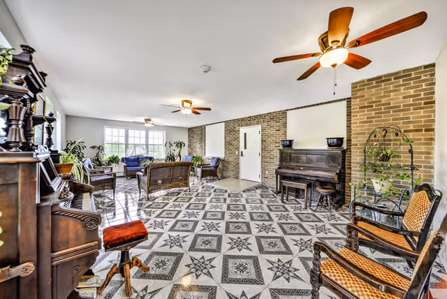 A spacious, well-lit common area with patterned tile flooring, brick walls, and multiple ceiling fans. The room is furnished with vintage wooden chairs, a piano, wicker seating, and various potted plants near large windows letting in natural light.