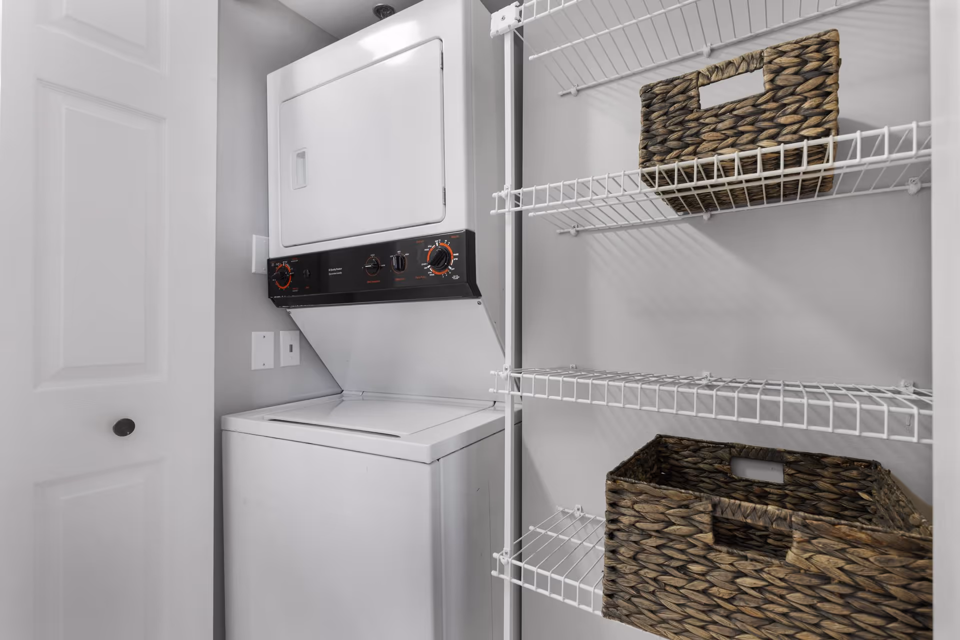 A small laundry area with a stacked white washer and dryer unit on the left and white wire shelving on the right holding two woven baskets.