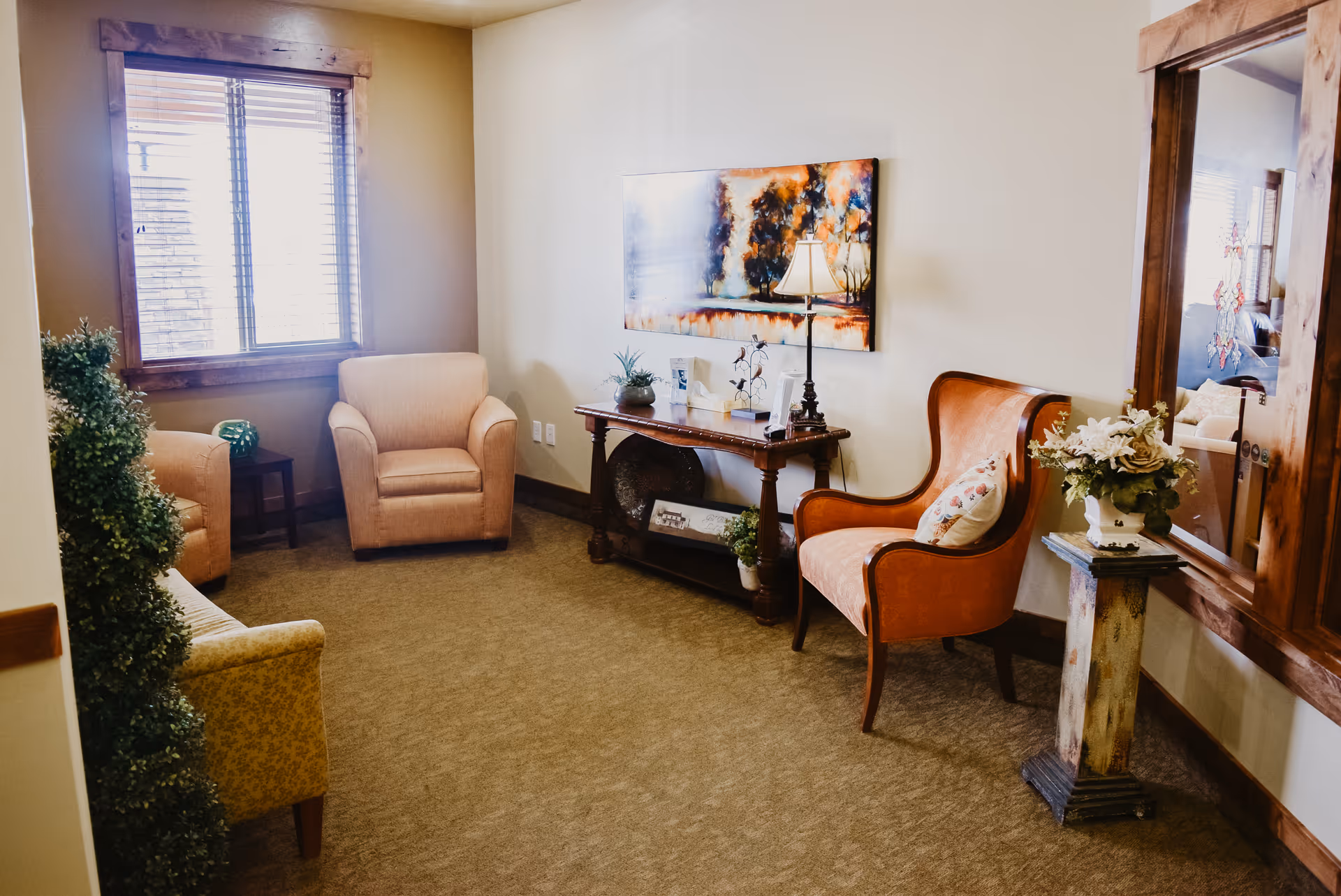 A cozy sitting area in Copper Summit Assisted Living featuring a beige armchair, an orange armchair with a decorative pillow, a small wooden table with a lamp and plants, a large mirror on the wall, and a window with wooden blinds letting in natural light.