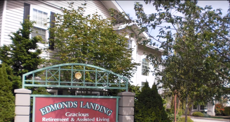 Exterior view of Edmonds Landing retirement and assisted living facility sign surrounded by trees and greenery, with part of the building visible in the background.