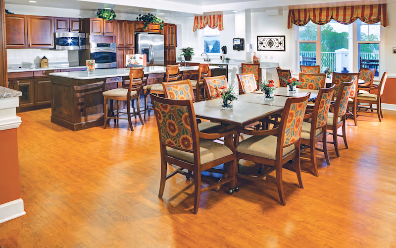 A spacious dining area with wooden flooring featuring multiple tables and chairs with floral-patterned upholstery. In the background, there is a kitchen area with wooden cabinets, a large island with bar stools, and stainless steel appliances. The room is well-lit with natural light coming through windows with striped valances.