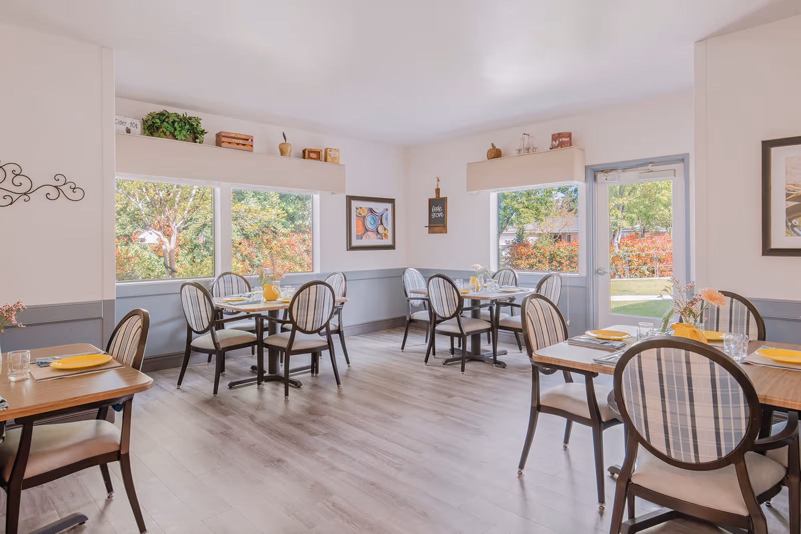 A bright and airy dining room with several wooden tables and chairs featuring plaid upholstery. The tables are set with yellow plates, glasses, and silverware. Large windows and a glass door provide a view of green trees and colorful bushes outside. The walls are painted white with a gray wainscoting, and there are decorative items and framed pictures on the walls and shelves.