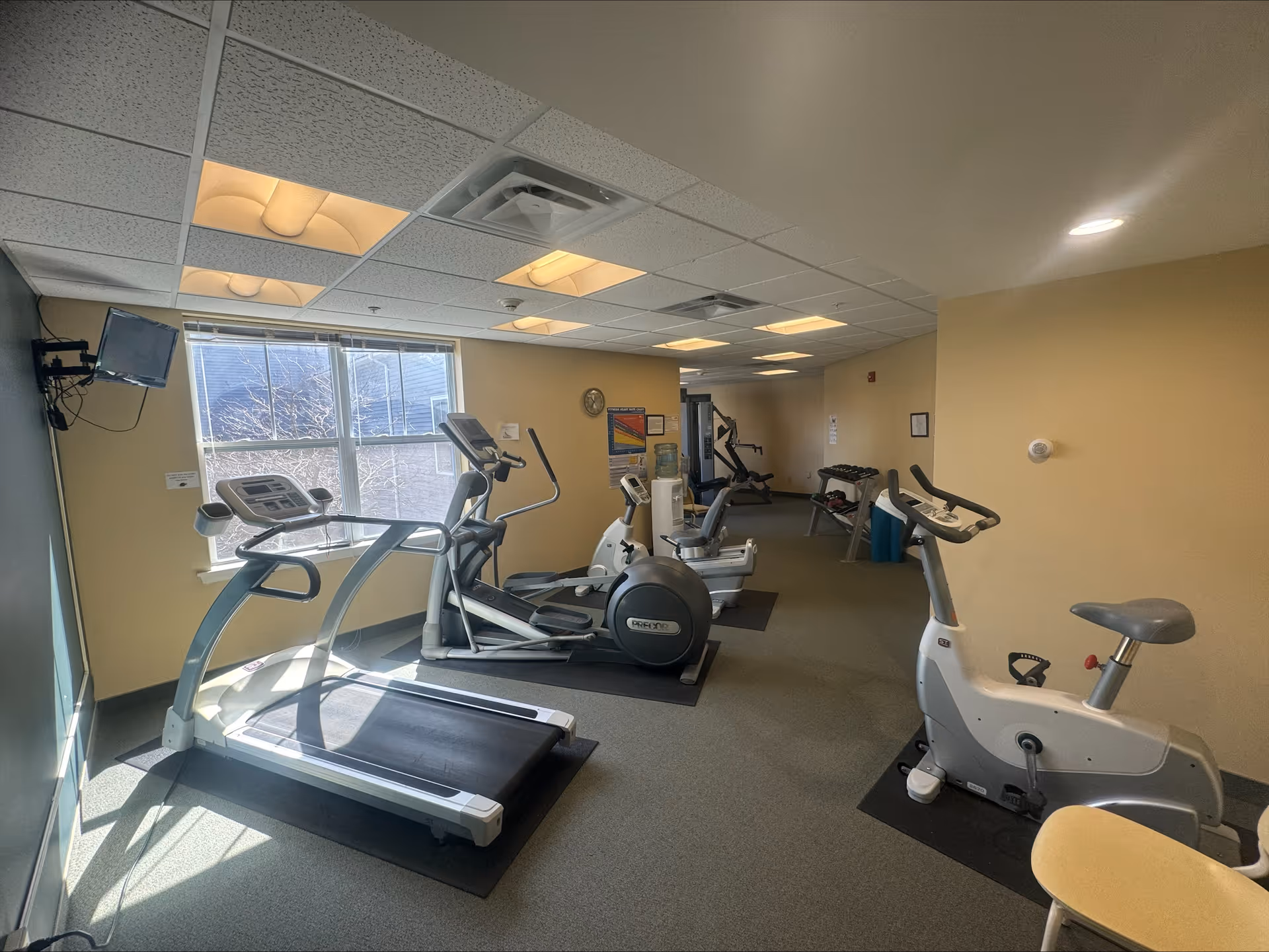 A small fitness room with exercise equipment including a treadmill, elliptical machine, stationary bike, and weight machine. There is a window letting in natural light, a wall-mounted TV, and a rack of dumbbells in the background. The walls are painted light yellow and the ceiling has recessed lighting.