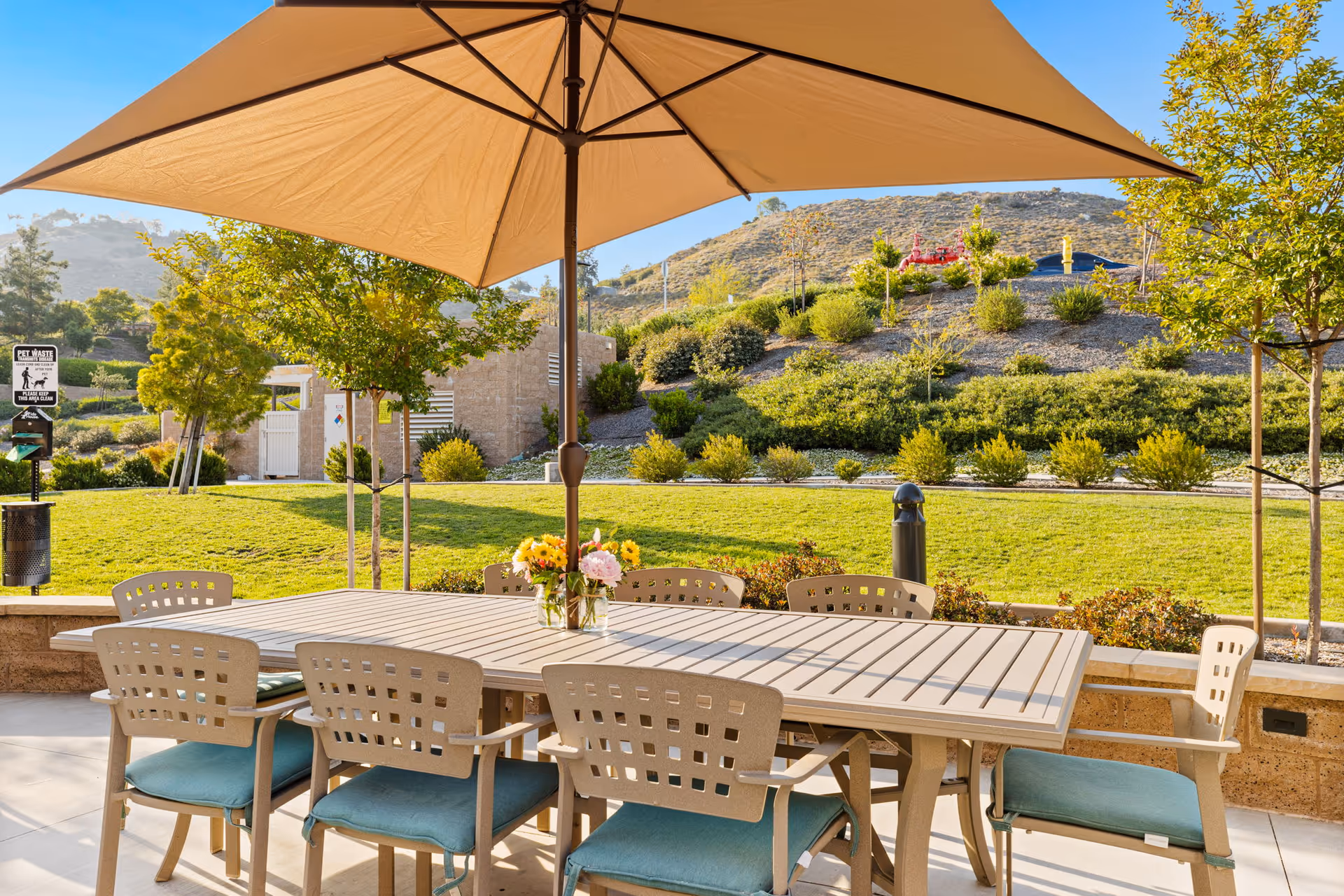 Outdoor patio area with a large rectangular table surrounded by eight chairs with blue cushions. A large beige umbrella provides shade over the table. In the background, there is a grassy area with trees, shrubs, and a hillside under a clear blue sky.