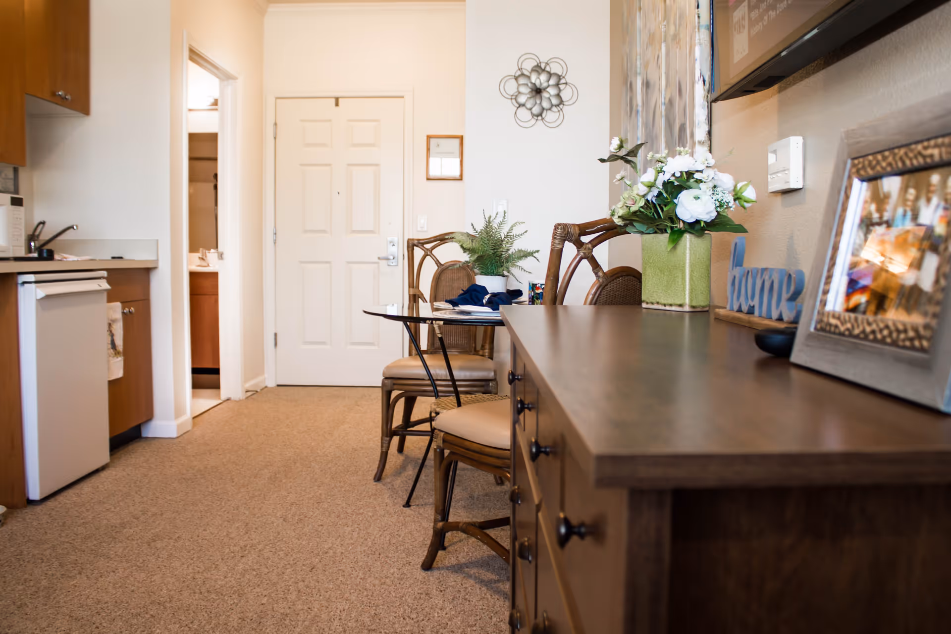 Interior view of a senior living facility apartment showing a small dining area with two chairs and a round table, a wooden dresser with decorative items including a flower vase and framed photos, a kitchenette with cabinets, a mini fridge, and a microwave, and a closed white door in the background.