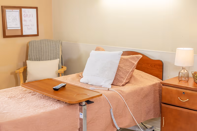 A cozy single hospital-style bed in a senior facility room with a bedside table, lamp, overbed tray with a remote, and a cushioned chair.