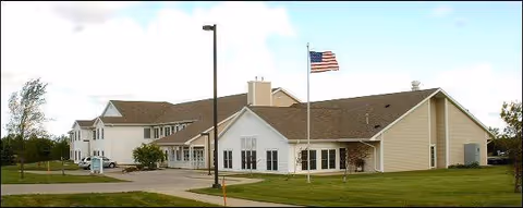 Exterior view of a single-story and two-story senior living facility building with beige siding and a brown roof. An American flag is flying on a flagpole in front of the building, and there is a paved driveway and green lawn surrounding the facility under a partly cloudy sky.