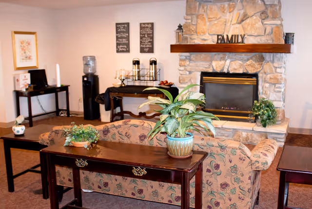 A cozy living room area with a floral-patterned sofa facing a stone fireplace. On the mantel above the fireplace is a decorative sign that reads 'FAMILY'. There are green plants on a wooden table behind the sofa and on the fireplace hearth. In the background, there is a water cooler, a small table with coffee dispensers, and framed wall art.