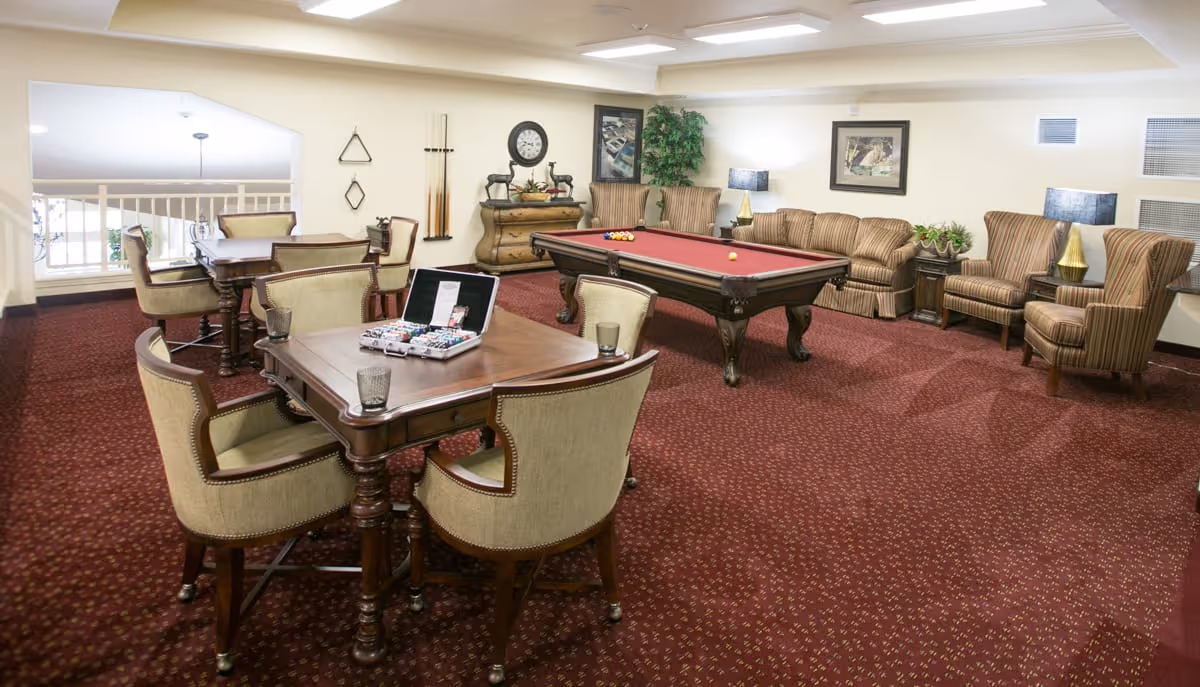 A cozy recreational room with a red carpeted floor featuring a pool table with balls set up, several upholstered chairs and sofas arranged along the walls, and two wooden tables with cushioned chairs. The room is decorated with framed artwork, a clock, a plant, and a few decorative items on a wooden dresser.