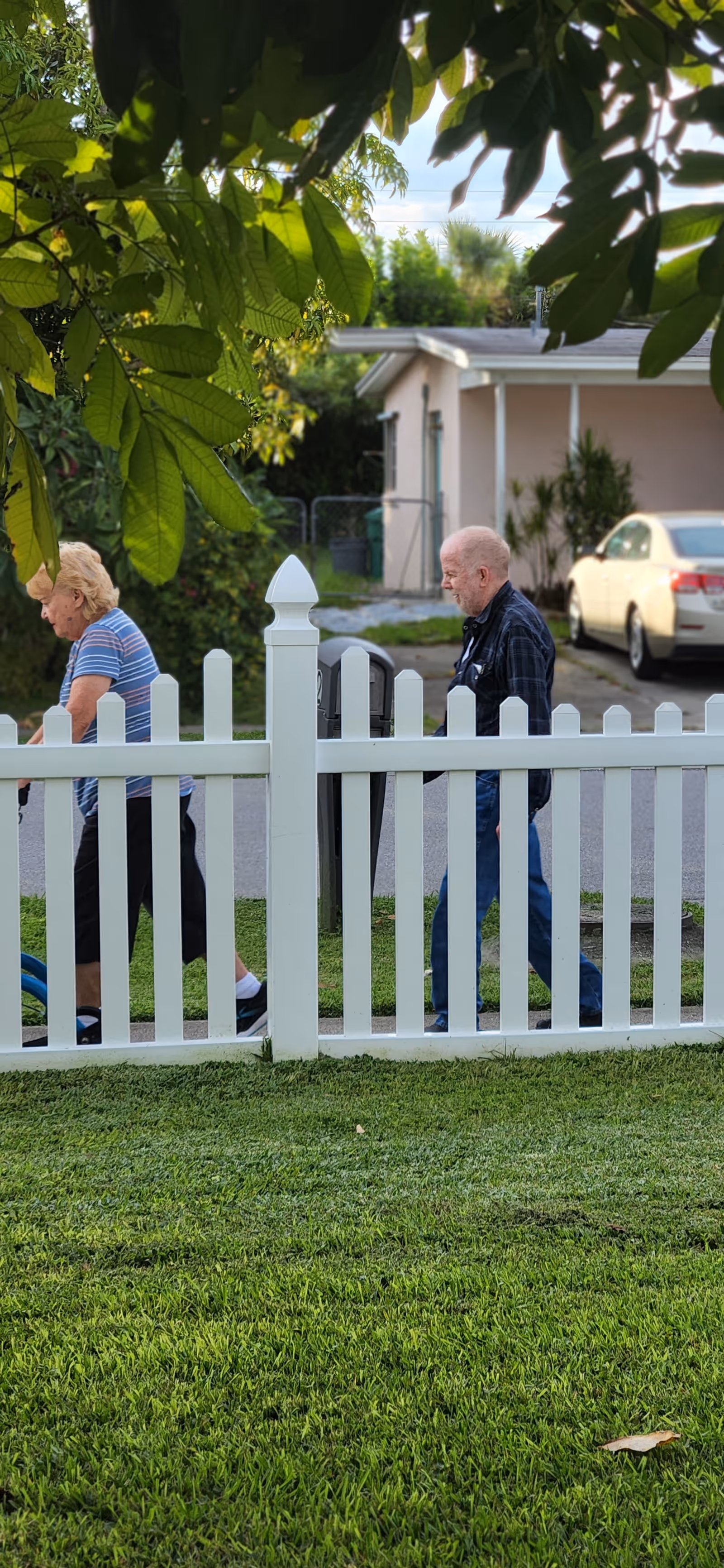 Two elderly individuals walking outside near a white picket fence with green grass and leafy trees overhead. In the background, there is a light-colored house and a parked car.