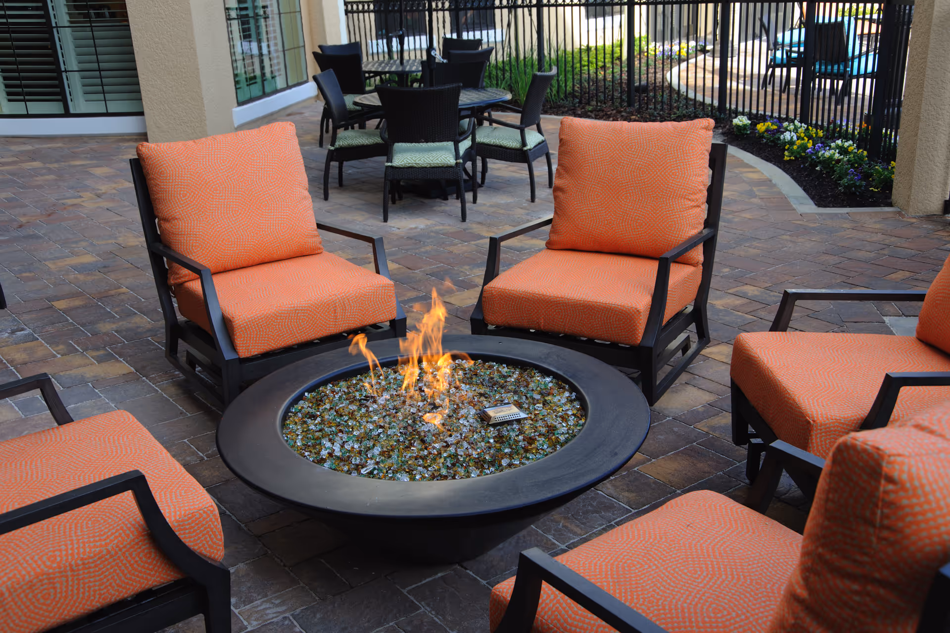 Outdoor patio with orange cushioned chairs arranged around a central glass-rock gas fire pit.
