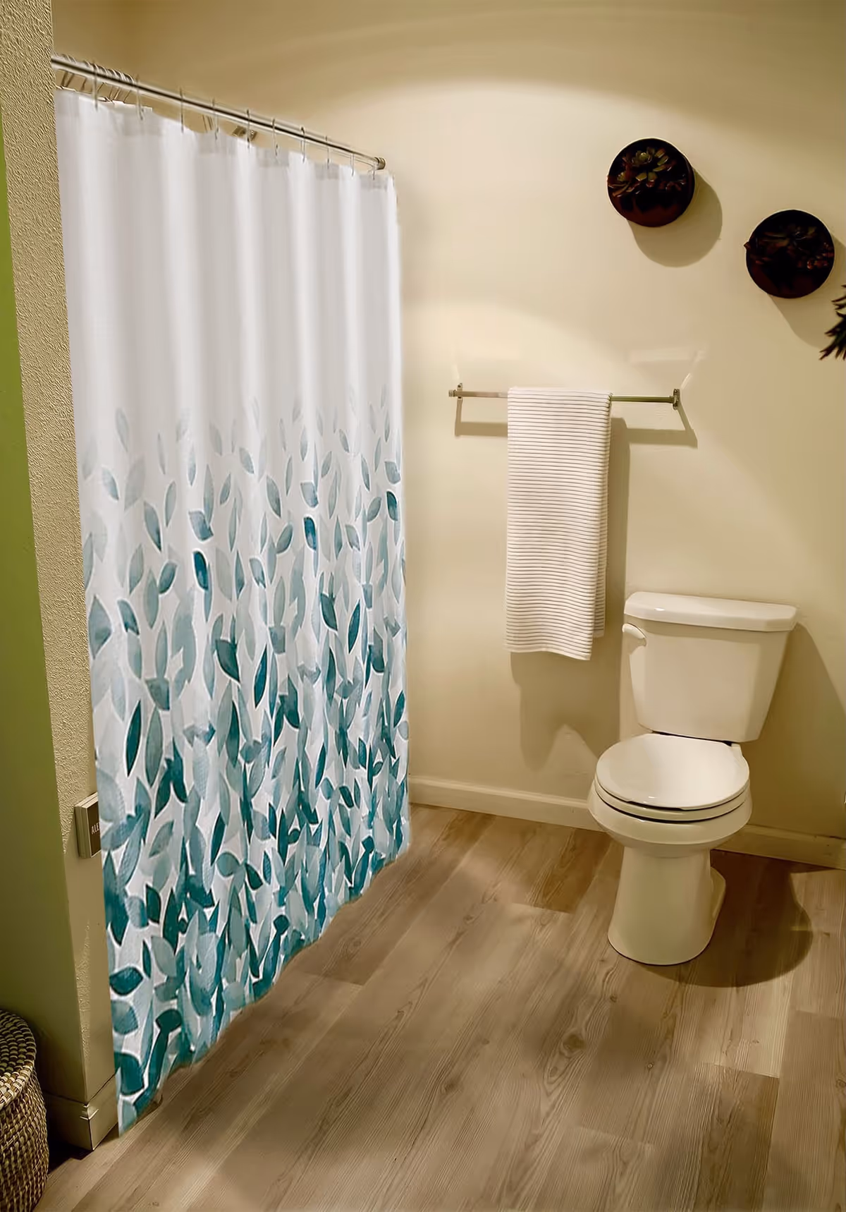 A bathroom with a white toilet, a towel rack holding a white towel, and a shower curtain decorated with green and blue leaf patterns. The floor is wooden, and two small round wall decorations are visible above the towel rack.