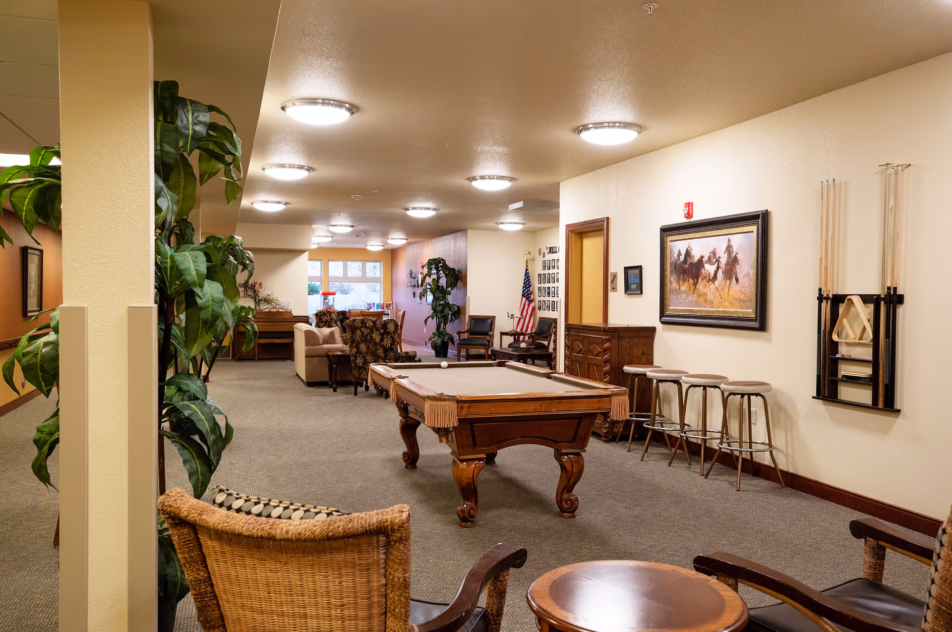 Interior view of a senior living facility common area featuring a pool table in the center, several chairs and stools, a painting of horses on the wall, an American flag, and plants. The space is well-lit with ceiling lights and has a carpeted floor.