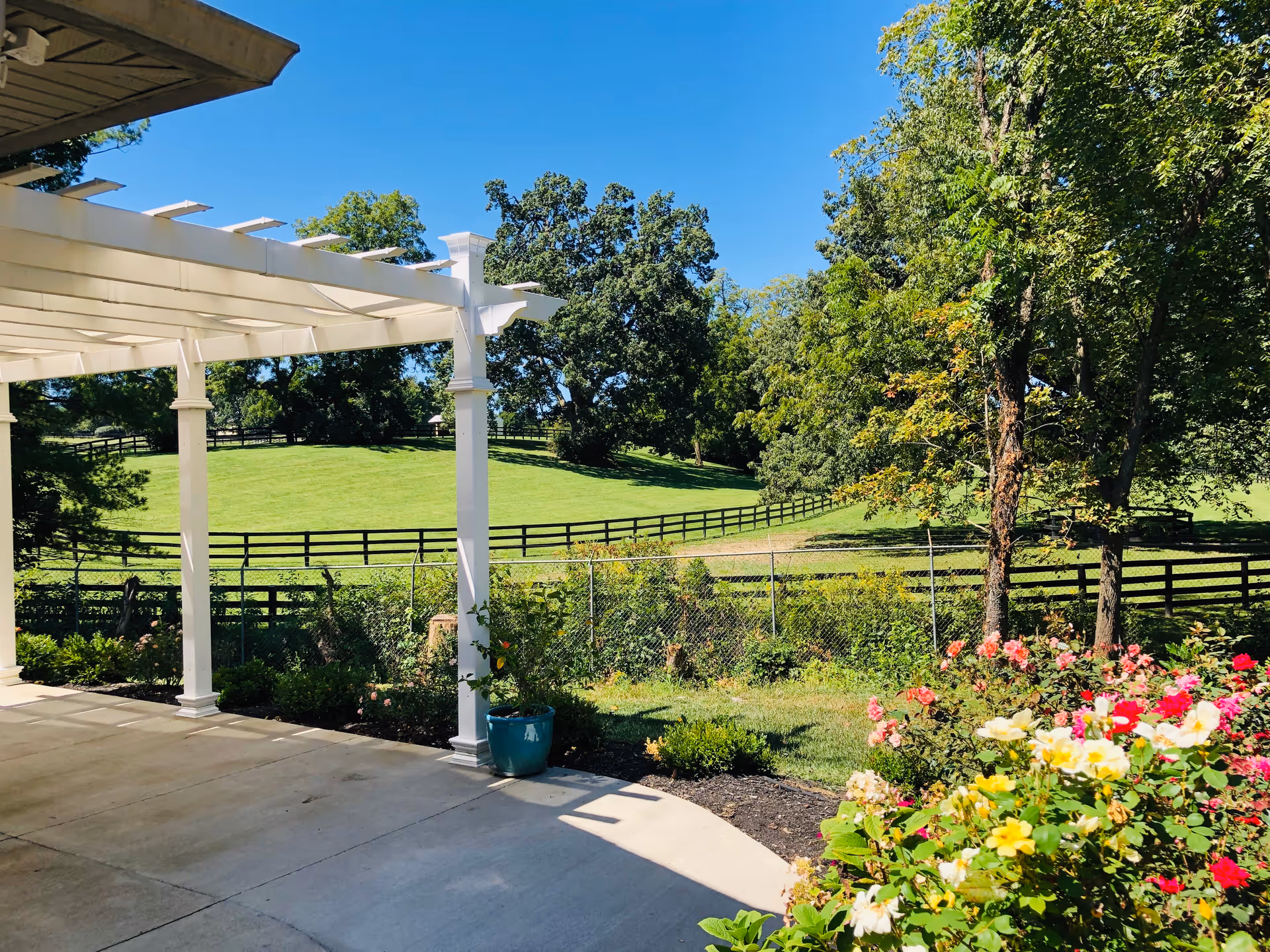 A sunny outdoor patio area with a white pergola structure, overlooking a green grassy field with a wooden fence and several trees. There are colorful flowers and shrubs planted along the edge of the patio.